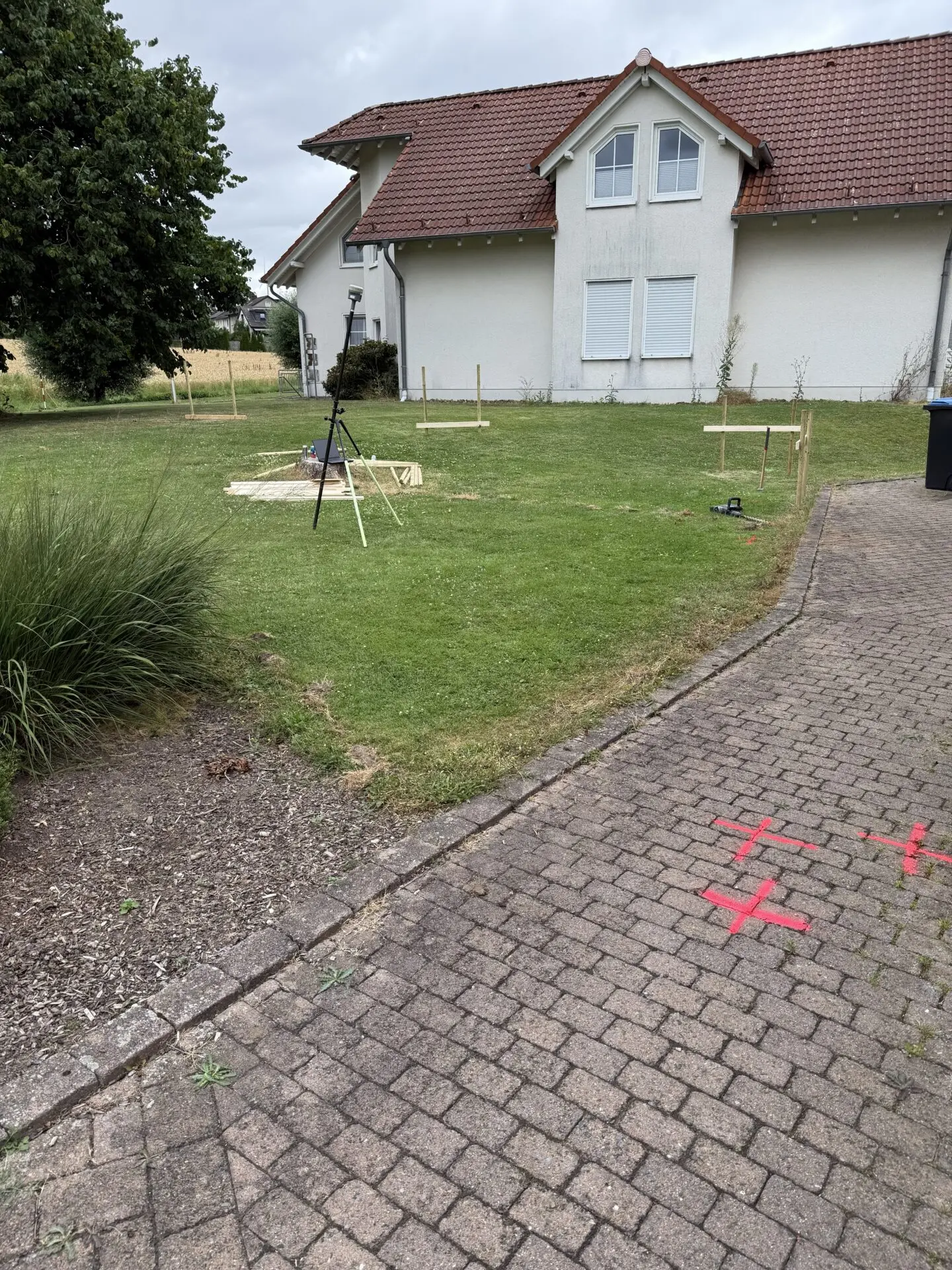 Vor einem weißen Haus mit rotem Dach sind auf der Wiese und dem Pflaster Vermessungsmarken für den Umbau zu sehen.