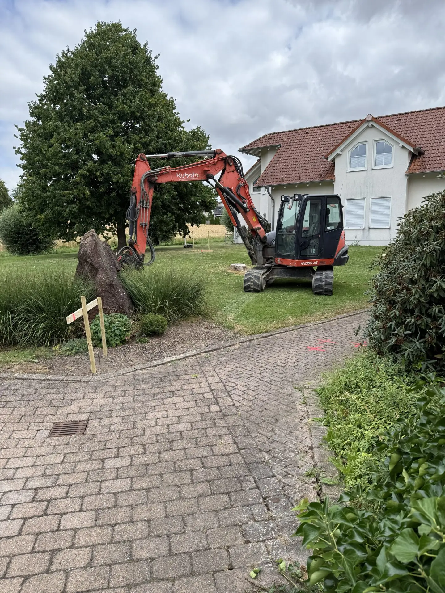 Ein orangefarbener Kubota-Bagger arbeitet im Garten vor einem weißen Haus mit rot gedecktem Dach unter wolkigem Himmel.